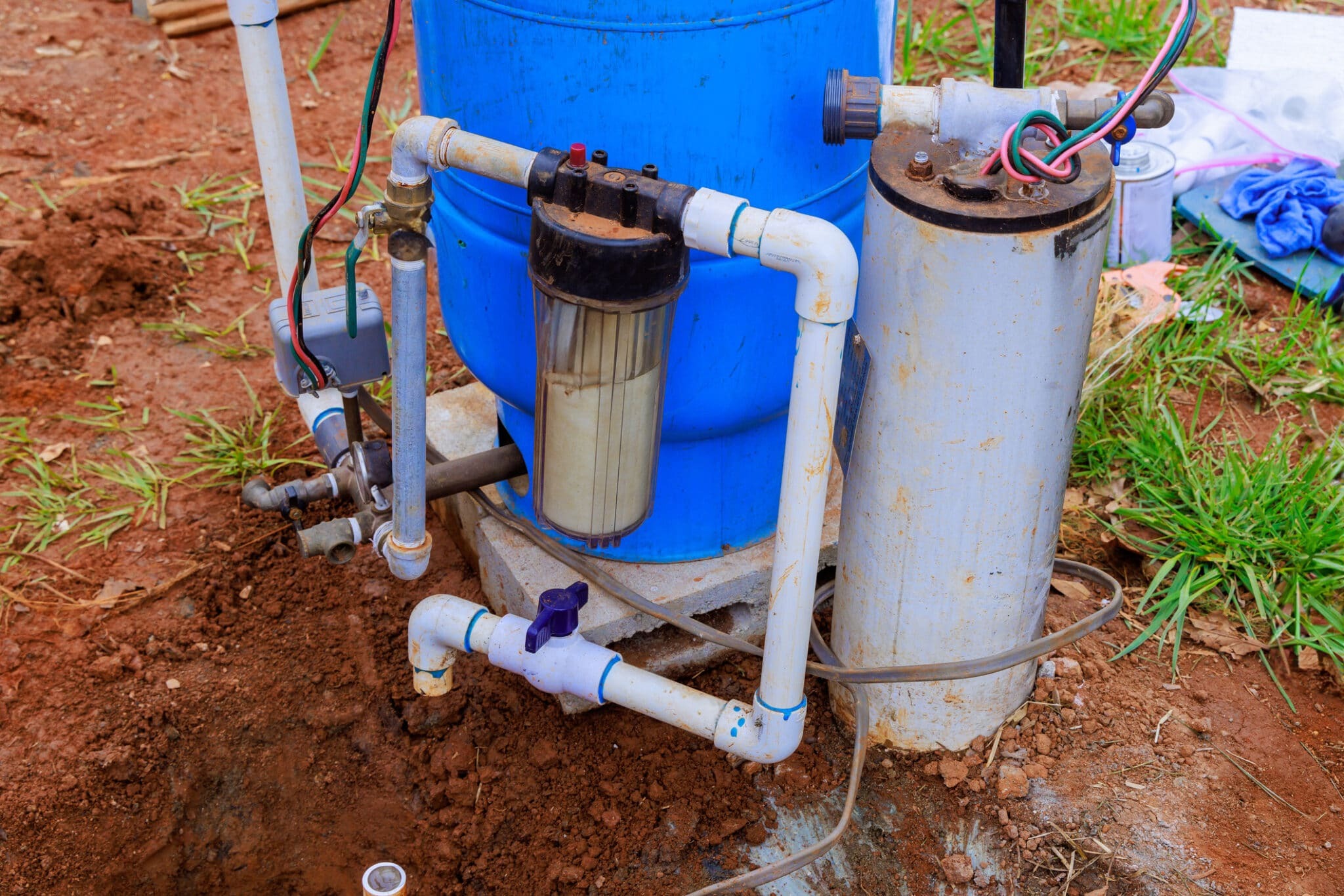 The equipment includes a blue tank, a filter, and pipes, set against a backdrop of dirt and grass.