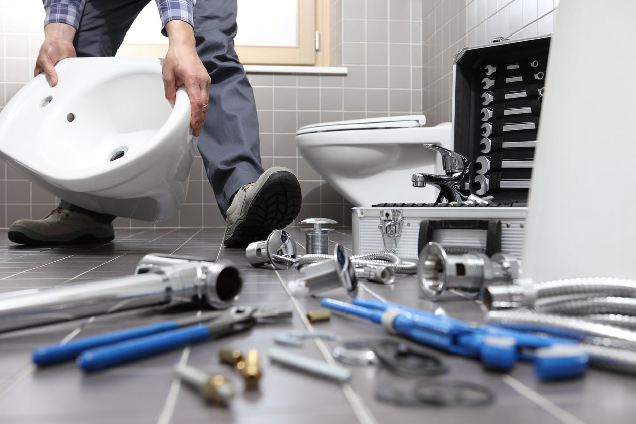 A plumber kneels in a tiled bathroom, holding a white sink above the floor.